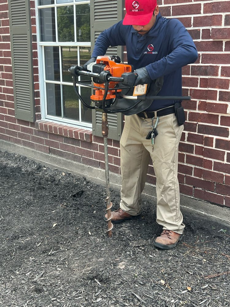 A Life After Bugs technician in a red cap using a power auger to drill next to a brick foundation for termite treatment