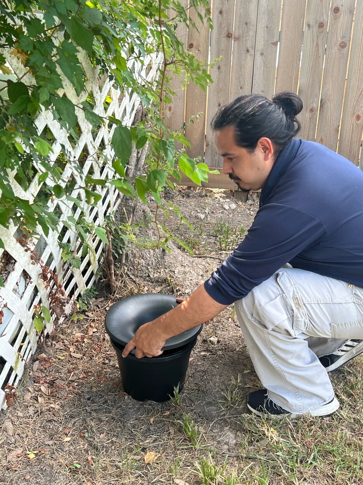 A technician crouching beside a black funnel-lid mosquito station installed in a backyard along a fence