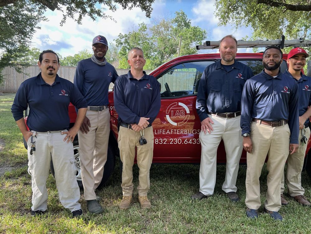 Life After Bugs technicians in navy uniforms standing on a lawn in front of a red company service truck